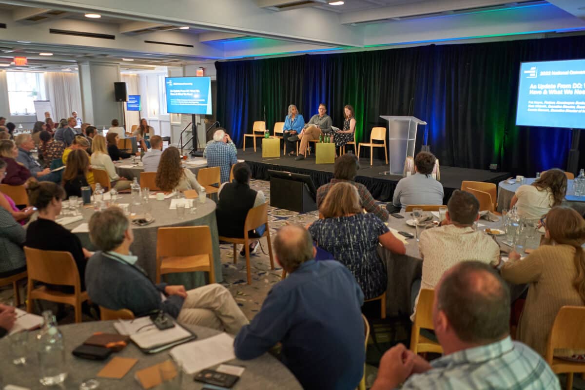 A panel of speakers on stage in front of a large crowd of guests at a conference.